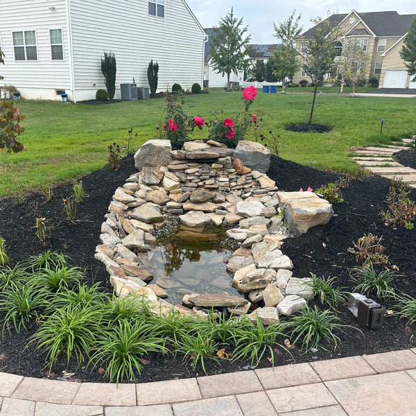 A custom-built natural stone waterfall and small garden pond surrounded by black mulch, featuring red flowering hibiscus and vibrant green lily turf (Liriope).